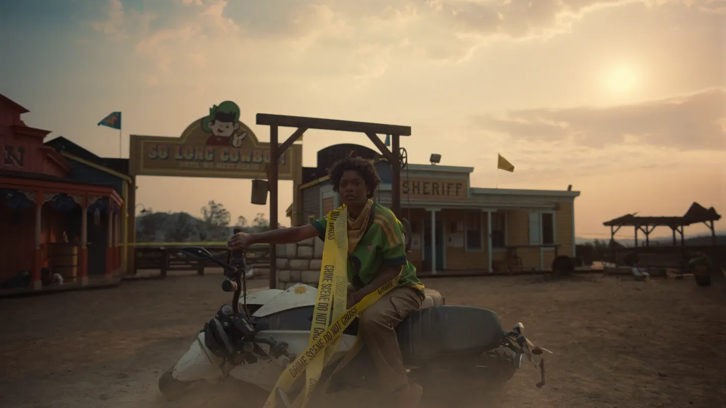 Nope — a woman sits astride a motorbike wrapped in crime-scene tape outside a western theme-park saloon at dusk. Additional music by Orlando Pérez Rosso.