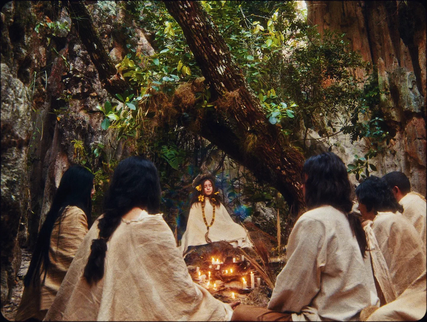 Simbiontes — a girl crowned with yellow flowers sits before a ring of candles as robed villagers look on in a Colombian forest. Original score by Orlando Pérez Rosso.